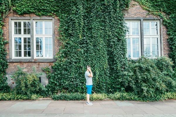 Man stands in front of building in ivy and listins music — Stock Photo, Image