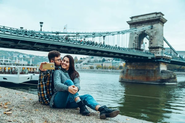 Szép cople ül a strandon, és figyelembe selfie bridge a háttérben — Stock Fotó