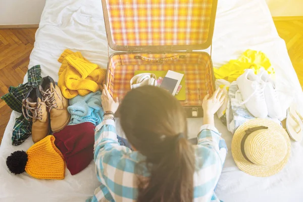 Žena si sedne na postel s kufrem a oblečení a okolí. cestovní koncept. rozhodnout, teplé nebo studené zemi jít. — Stock fotografie