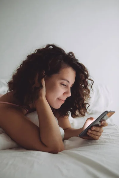 Jong mooi vrouw met krullend haar in bed met telefoon — Stockfoto