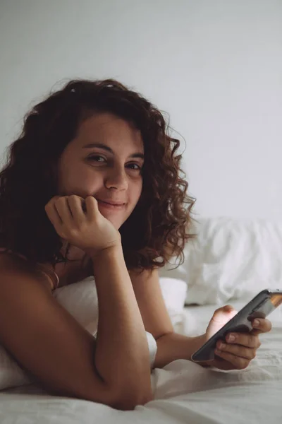 Fiatal csinos nő göndör haj az ágyban telefonnal — Stock Fotó
