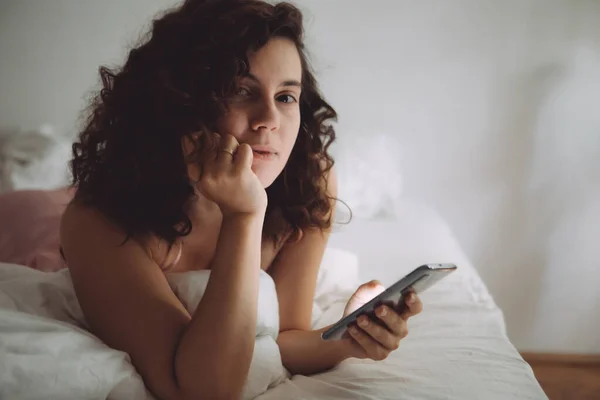 Joven bonita mujer con pelo rizado en la cama con teléfono —  Fotos de Stock