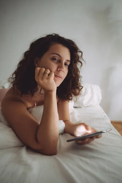 Jovem mulher bonita com cabelo encaracolado na cama com telefone — Fotografia de Stock