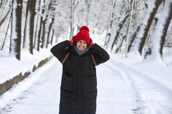 Fiatal csinos mosolygós nő télikabátban piros kalap a városi erdő parkban — Stock Fotó
