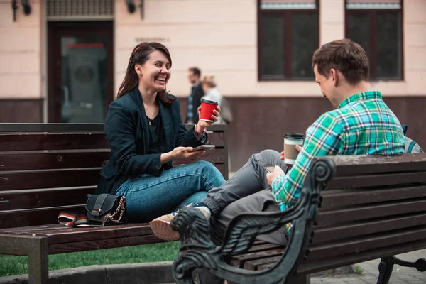 Смеющийся мужчина с женщиной, сидящей на скамейке и пьющей кофе-серфинг в интернете. городской образ жизни — стоковое фото
