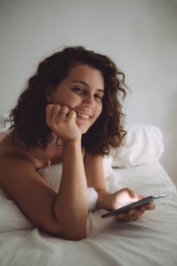 Yatakta kıvırcık saçlı, telefonlu genç ve güzel bir kadın.