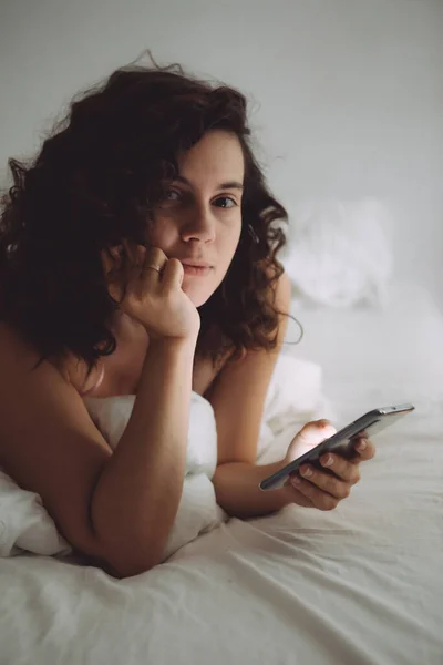 Young pretty woman with curly hair in bed with phone — Stock Photo, Image