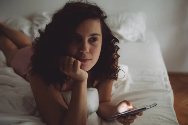 Young pretty woman with curly hair in bed with phone — Stock Photo, Image