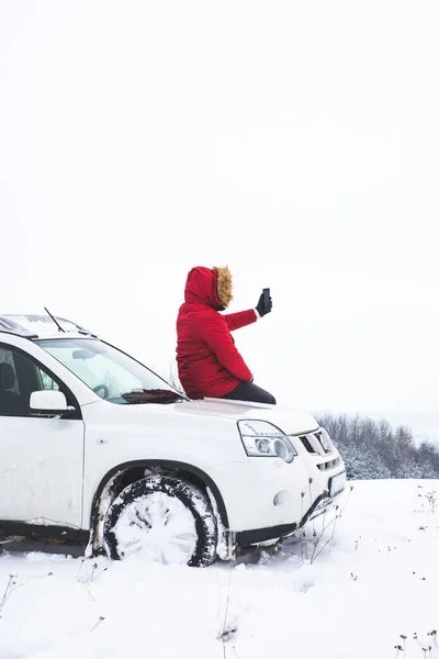Hombre sentado en la capucha del coche y fotografía de la hermosa vista de invierno en su teléfono — Foto de Stock
