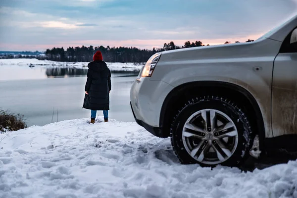 Fiatal felnőtt nő téli ruhák piros kalap bubo áll közel SUV autó folyó mellett gyönyörű kilátással — Stock Fotó