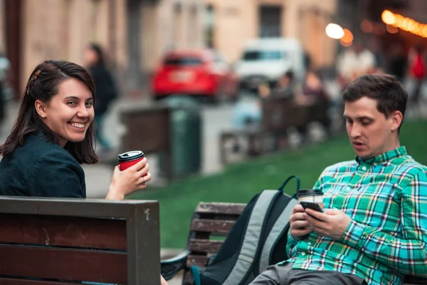 Smiling young woman drinking coffee while sitting on city bench man surfing internet with smartphone — Stock Photo, Image