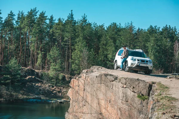 Joven en el borde con hermosa vista del lago cerca de coche suv blanco — Foto de Stock