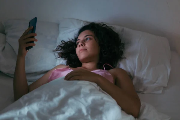 晚上带着手机躺在床上的年轻女子 — 图库照片
