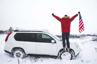 Tepenin tepesinde duran bir adam elinde ABD bayrağı ile SUV arabasının yanında güzel bir kış manzarası arıyor.