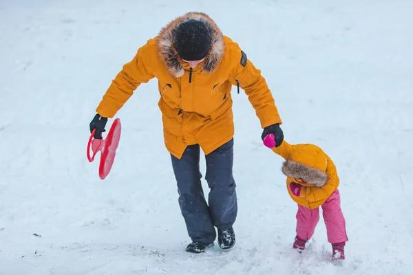 雪に覆われた丘を滑り降りる黄色い冬のコートを着た娘と — ストック写真