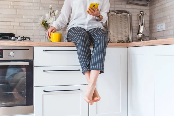 Mulher sentada na mesa da cozinha com telefone e caneca amarela — Fotografia de Stock