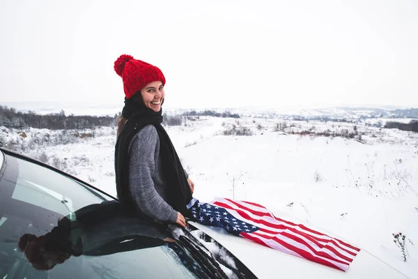 Νεαρή ενήλικη γυναίκα ταξιδιώτη κάθεται στο καπό του SUV αυτοκίνητο με σημαία ΗΠΑ — Φωτογραφία Αρχείου