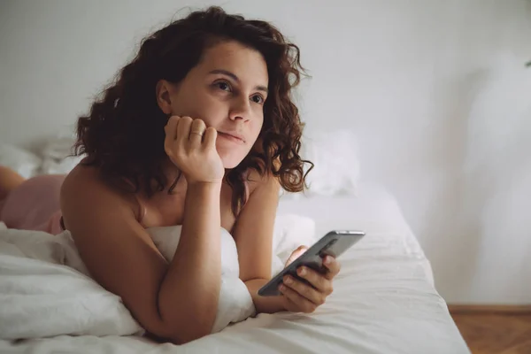 Young pretty woman with curly hair in bed with phone — Stock Photo, Image