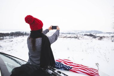 Araç kaputunda oturan genç, yetişkin bir kadın gezgin. Amerikan bayrağıyla telefonunda fotoğraf çekiyor.