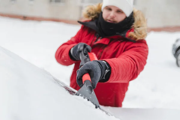 Man i röd vinterrock med päls huva rengöring bil efter snöstorm — Stockfoto
