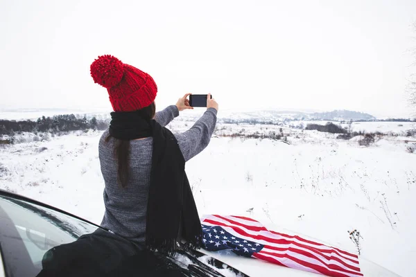 Νεαρή ενήλικη γυναίκα ταξιδιώτη κάθεται στο καπό του SUV αυτοκίνητο με σημαία ΗΠΑ λήψη φωτογραφιών στο τηλέφωνό της — Φωτογραφία Αρχείου