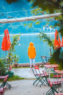 Sarı yağmurluklu kadın göle bakıyor. Kafe masaları ön tarafta.