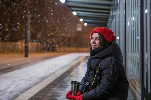 Mujer en traje de invierno con sombrero rojo sentado en la estación de autobuses esperando el transporte público en invierno noche de niebla — Foto de Stock