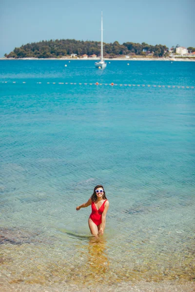 Woman in red swimsuit in white sunglasses standing in sea. copy space. summer vacation. yacht on background — Stock Photo, Image