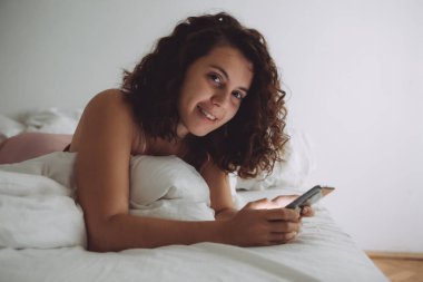 Yatakta kıvırcık saçlı, telefonlu genç ve güzel bir kadın.