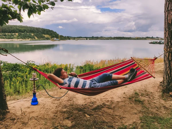 Pria berbaring di tempat tidur gantung merokok hookah melihat danau pantai — Stok Foto