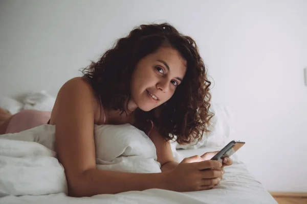 फोन के साथ बिस्तर में घुमावदार बालों वाली युवा सुंदर महिला — स्टॉक फ़ोटो, इमेज