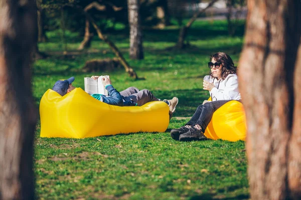 Coppia che posa su materasso gonfiabile giallo nel parco cittadino. leggere un libro. bere frullati — Foto Stock