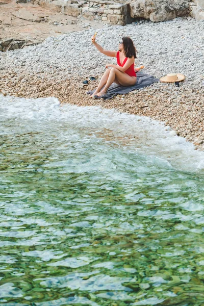 Giovane bella donna in costume da bagno rosso seduta sulla spiaggia rocciosa con telefono scattare foto selfie — Foto Stock