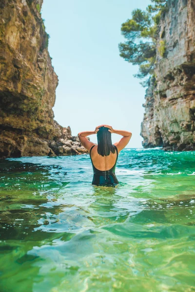 身穿黑色泳衣的年轻成年女子走在岩石悬崖之间的海面上 — 图库照片