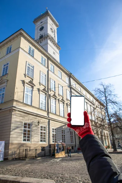 Mano de mujer en guantes rojos sosteniendo el teléfono con pantalla blanca Ayuntamiento de Lviv en el fondo — Foto de Stock