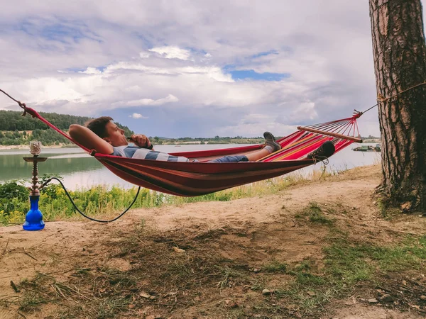 Muž ležící v houpací síti kouření hookah při pohledu na pláž jezera — Stock fotografie