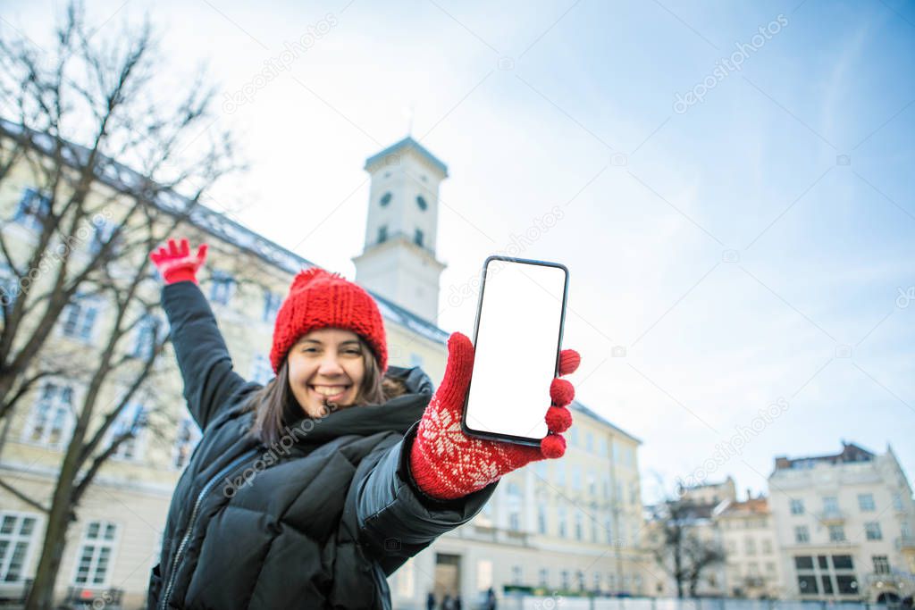 man in red winter hat holding phone with white empty blank screen european city hall on background