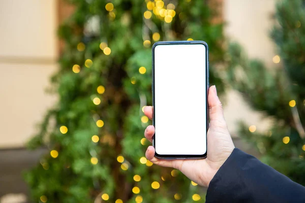 Γυναίκα χέρι κρατώντας τηλέφωνο με λευκή οθόνη χριστουγεννιάτικο δέντρο στο παρασκήνιο — Φωτογραφία Αρχείου
