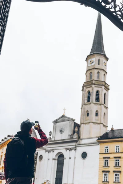 Férfi turista esőkabátban hátizsákkal nézi Szent Mihály templom fényképezés a telefonon — Stock Fotó