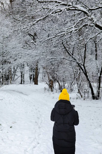 Woman in black winter coat and yellow hat walking by snowed city park — Stock Photo, Image