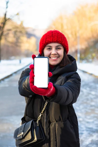 Young smiling woman in winter outfit holding phone with white blank empty screen — Stock Photo, Image