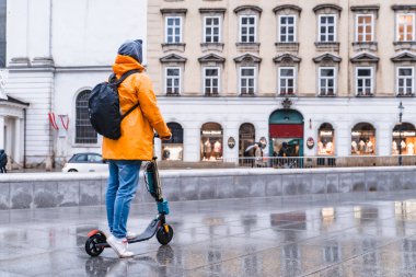 Sarı yağmurluklu bir adam elektrikli scooter ile şehir sokağının yanından geçiyor.
