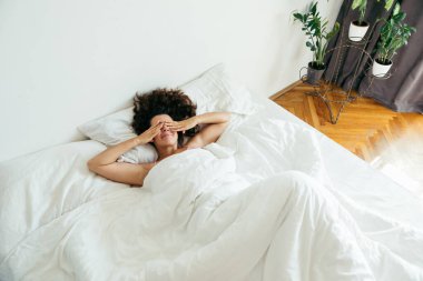 Genç ve güzel bir kadın beyaz çarşaflı parlak bir ışıkla yatakta.