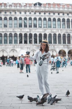 Beyaz giysili, hasır şapkalı kadın Venedik Meydanı 'nda güvercinlerle eğleniyor.