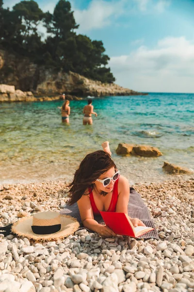 Frau in rotem Badeanzug liegt auf Decke am Strand und liest Buch — Stockfoto