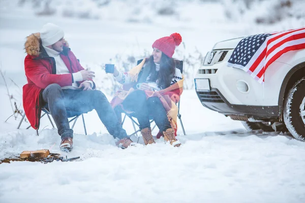 Couple sitting in chairs near campfire talking. suv car with usa flag on the hood — Stock Photo, Image