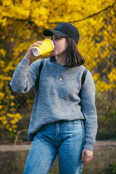 戴着帽子的年轻时尚女人在户外用黄色纸杯喝咖啡 背景的黄色开花灌木 — 图库照片