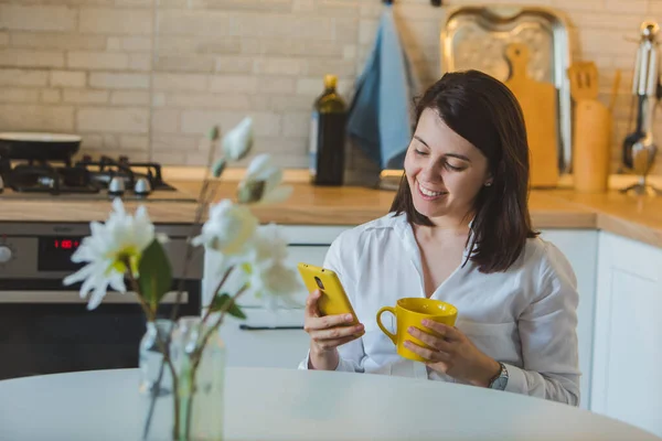 Žena Pije Čaj Žlutého Hrnku Kuchyni Surfování Internetu Telefonu — Stock fotografie