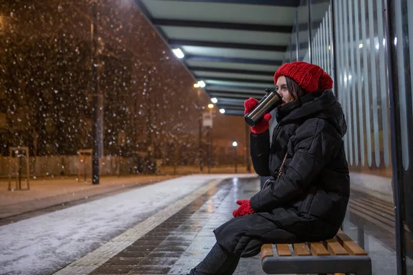 woman in winter outfit with red hat sitting at bus station waiting for public transport at winter snowing night copy space