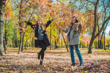 two girlfriends fooling around with yellow leaves at city park autumn fashion